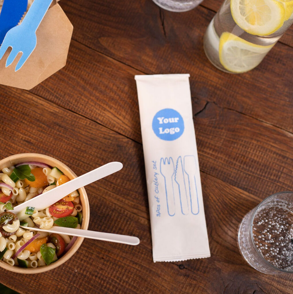 On a dark wooden table, a three-piece eco-friendly cutlery set in a branded wrapper is displayed next to a salad.